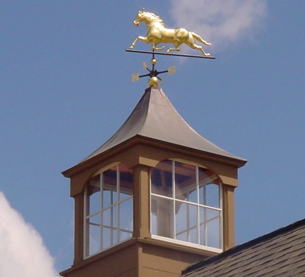 Gold horse weathervane mounted on a vinyl cupola for barns, featuring a windowed design and copper roof by Country Cupolas and Weathervanes.